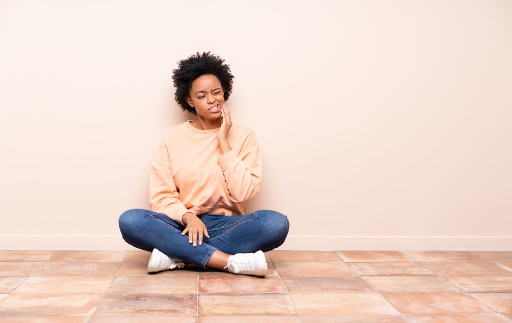 woman sitting on the floor holding her jaw, tmj pain​​​​​​​