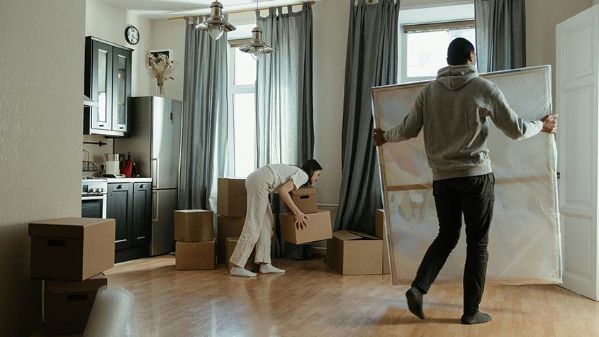 Two people moving boxes and a large wrapped item in a bright room during a relocation.