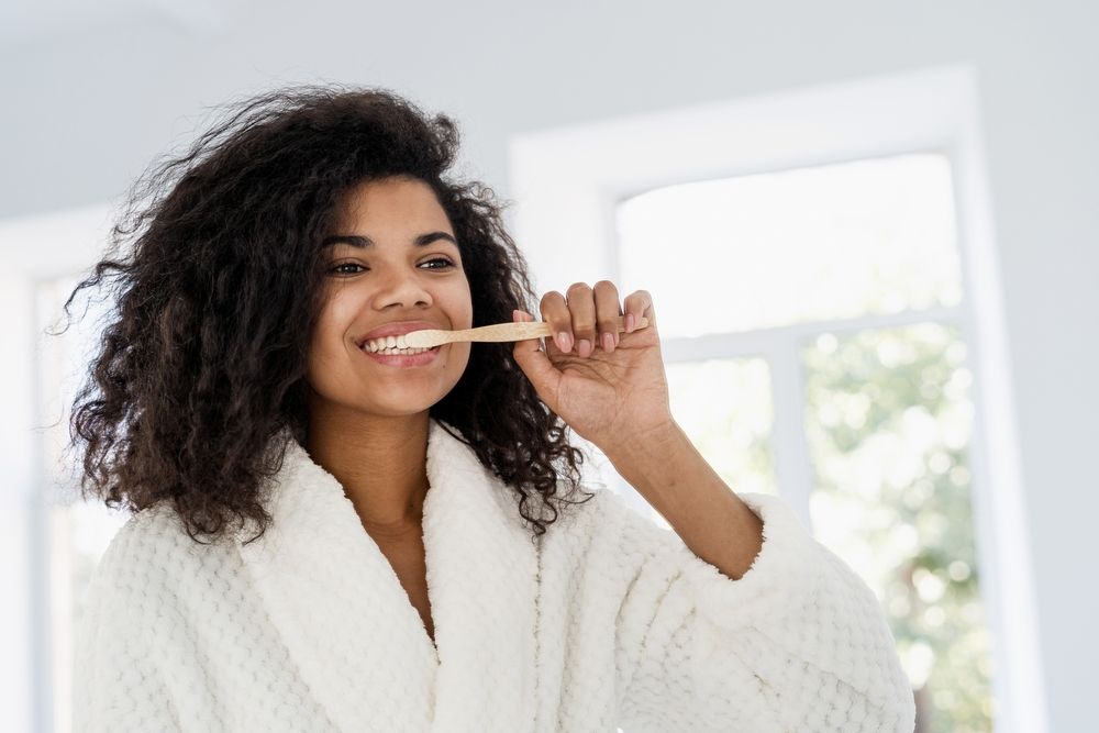 Personal oral hygiene and dental care concept. Portrait of happy young woman cleanse sensitive teeth with a bamboo brush and whitening toothpaste. Female in bathrobe looking at mirror
