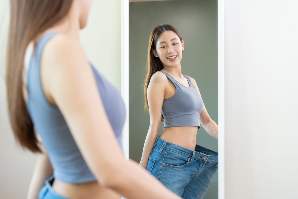 a woman who has lost weight happily looks at herself in the mirror wearing her old larger-fitting jeans