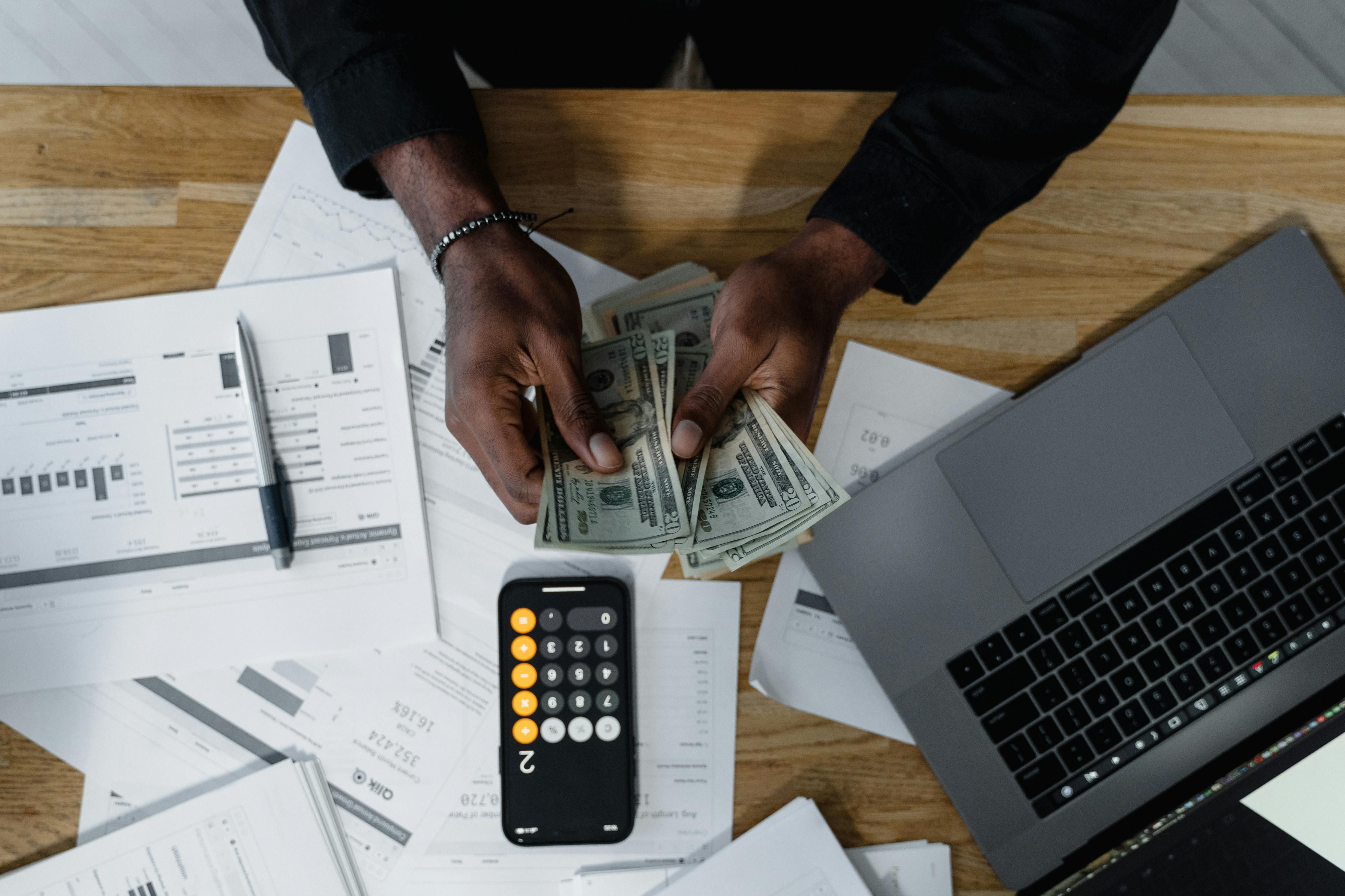 Man sitting at a desk and counting money, surrounded by documents, a laptop, and a phone