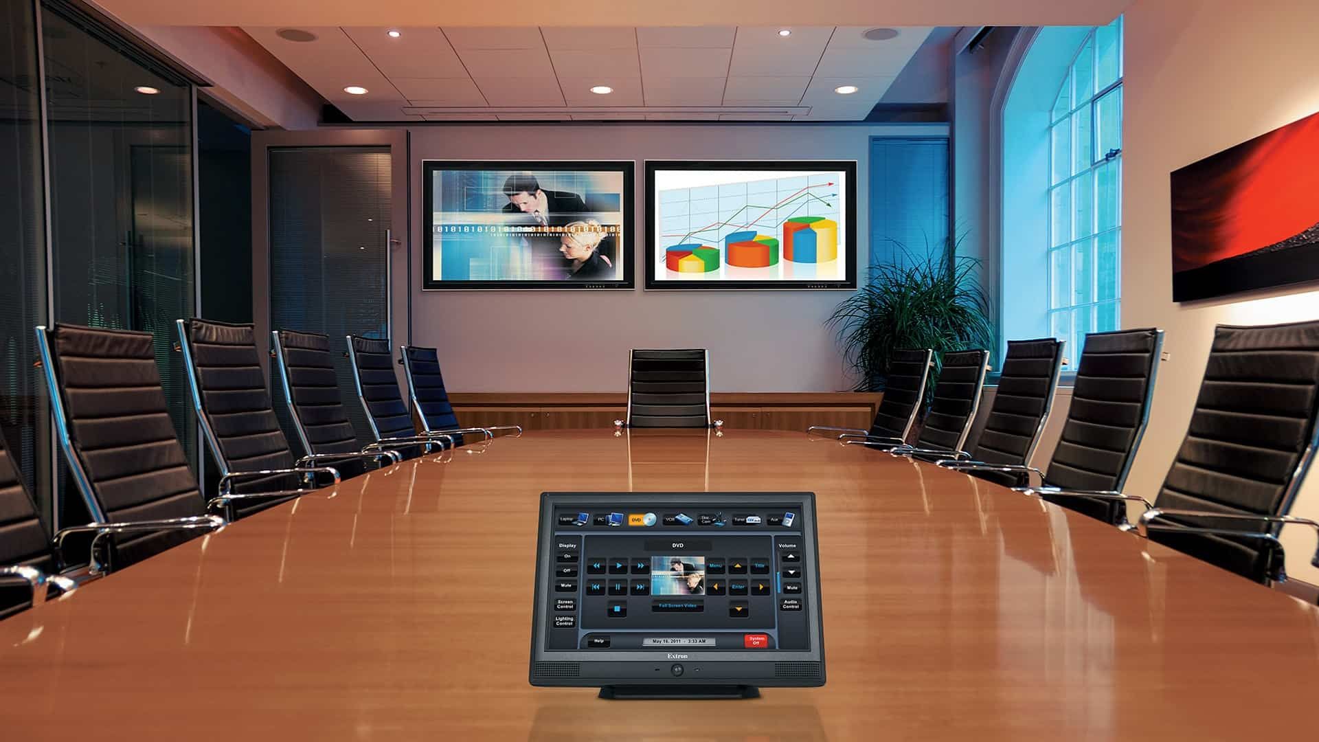 Picture of a board meeting room. There are more than 10 black chairs around a long table with a table controlling two TVs at the end of the room